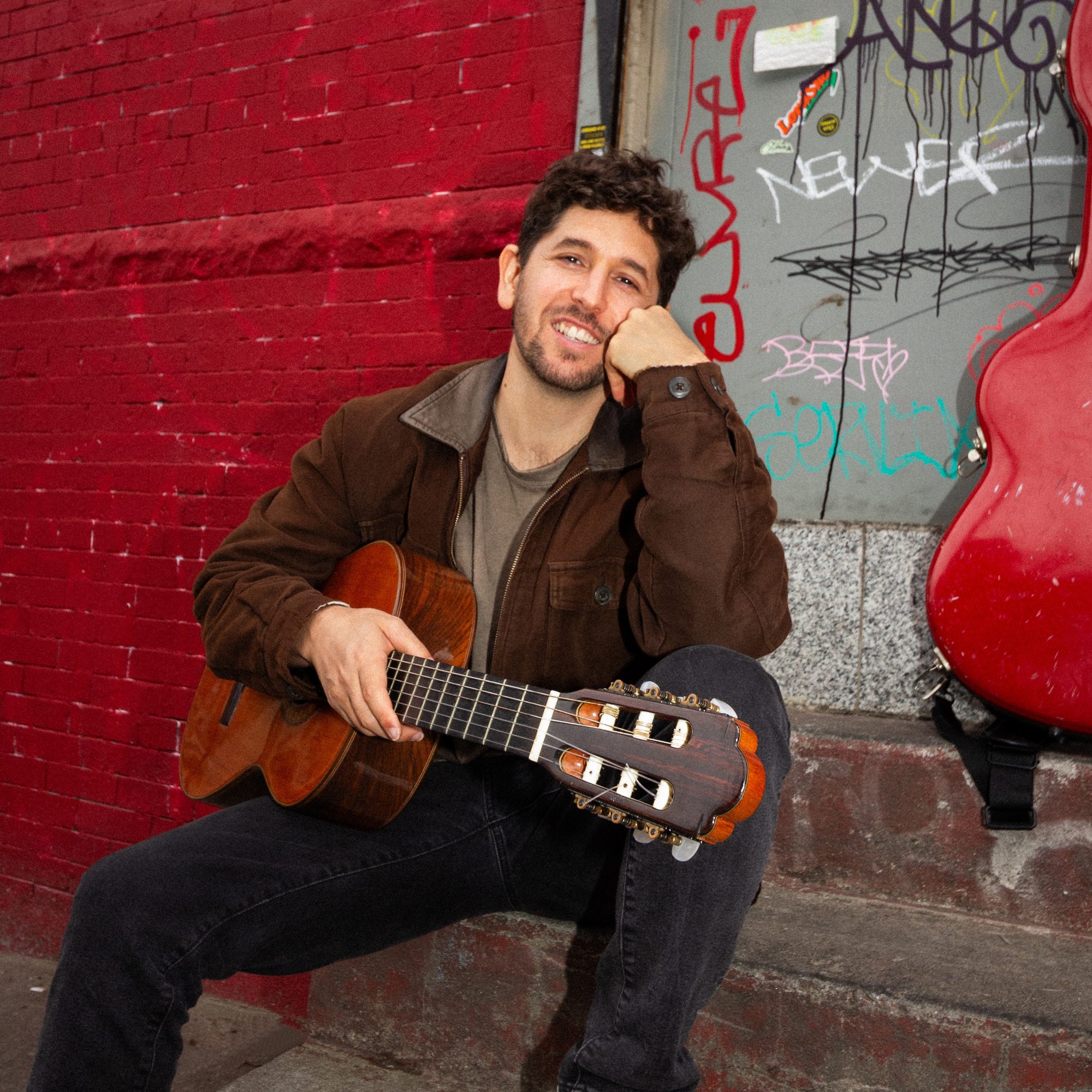 Tal Mashiach smiling with his classical guitar by a red brick wall